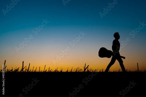 Silhouette of a musician holding a guitar and walking along the horizon at sunset © blumbaker