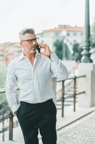 Businessman in good mood is talking on cellphone on his way to office. Handsome cheerful manager in formal suit walking enjoying getting to job in morning by foot having mobile conversation