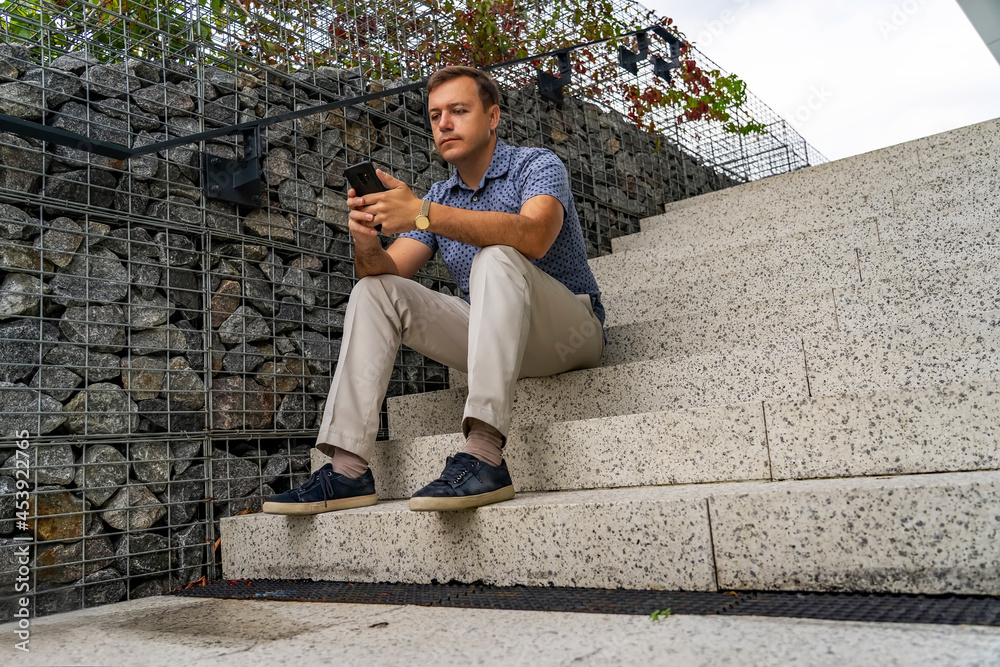 Portrait of handsome man sitting on stairs outdoors, using mobile phone and thinking. Business man checking e-mail