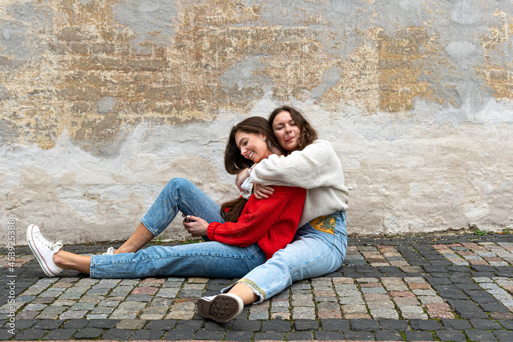 two young women hugged sitting on a paved sidewalk by an old wall