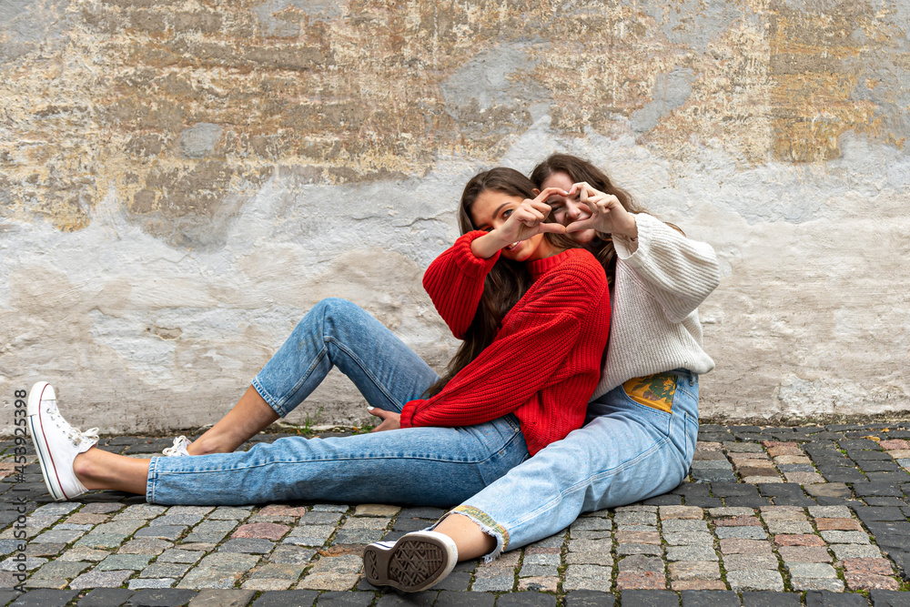 two young women hugged sitting on a paved sidewalk by an old wall