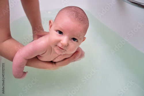 Mom trains swimming with a happy infant baby boy in a home bath. A smiling child swims n the bathroom in the hands of his mother
