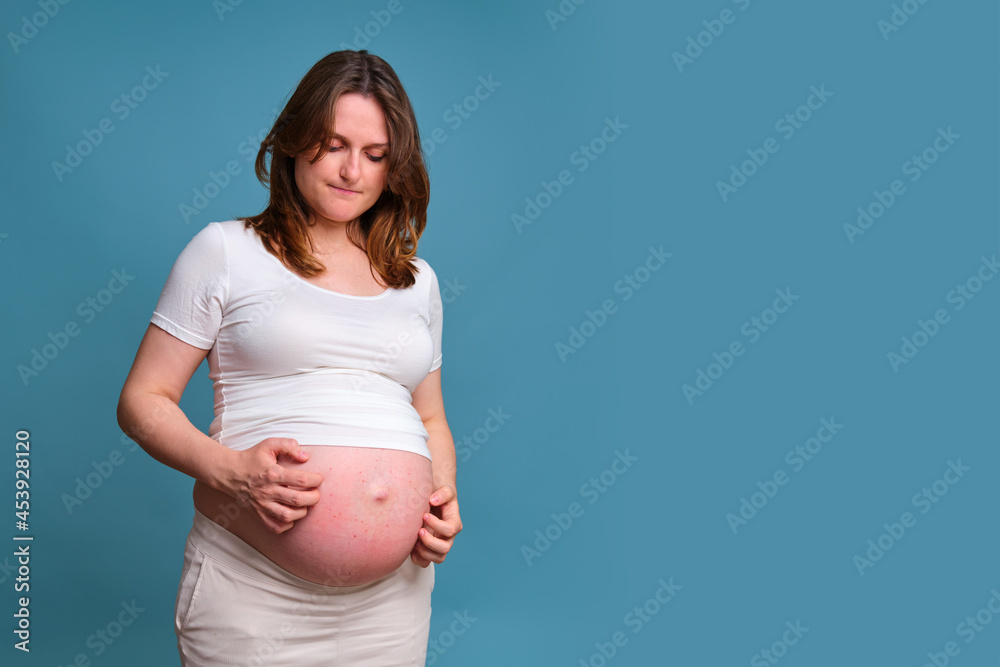 Portrait of a pregnant woman scratching her stomach, blue background Portrait of a pregnant woman scratching her stomach, blue background
