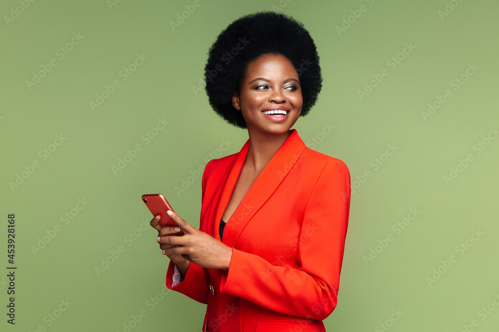 Successful african businesswoman hold smartphone look aside with confident happy smile. Afro american young entrepreneur female in luxury red suit with mobile phone isolated over green studio wall - obrazy, fototapety, plakaty 