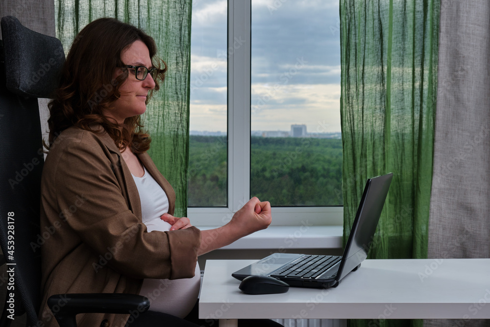 Pregnant woman with laptop in home office shows fist Pregnant woman with laptop in home office shows fist