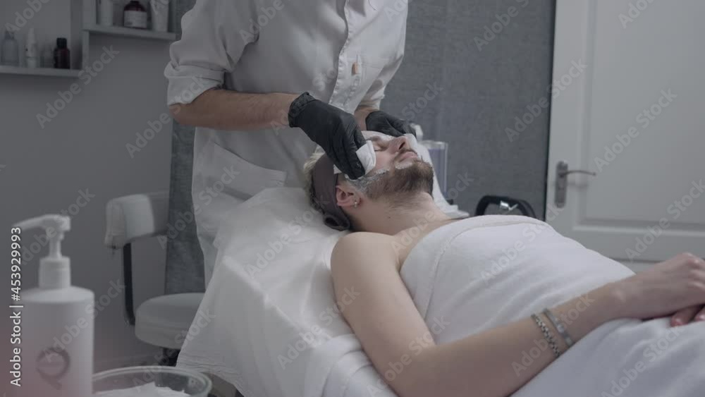 Beauty therapist wiping serum moisturizer from face of young man after LED light procedure in beauty parlor. Unrecognizable man serving male Caucasian client in spa salon indoors. Slow motion