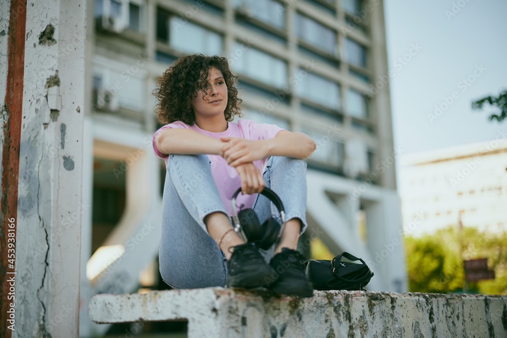 An attractive young woman with curly hair sitting outdoors and holding headphones in her hands. Urban living concept. An attractive young woman with curly hair sitting outdoors and holding headphones in her hands. Urban living concept.