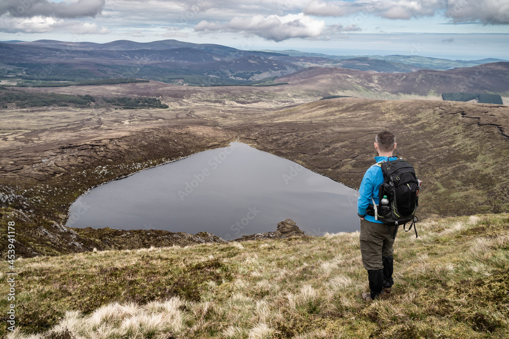 Hiker is standing on the top of the mountain with hands in the pockets. Back view.. Heart shaped Lake Ouler Tonelagee Mountain, Wicklow County, Ireland.