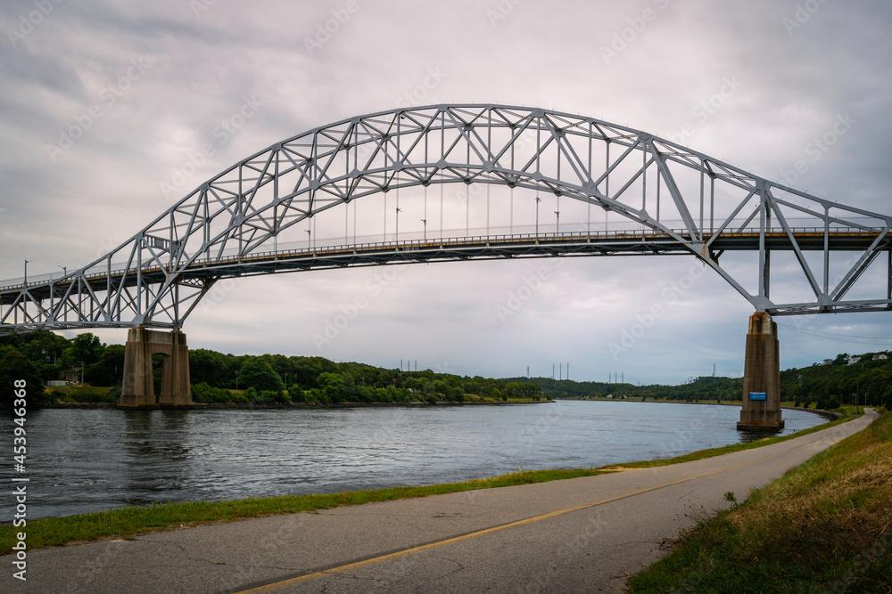Sagamore Bridge over Cape Cod Canal and bikeways along the riverbank. Dramatic cloudscape over the arching bridge over the river. 