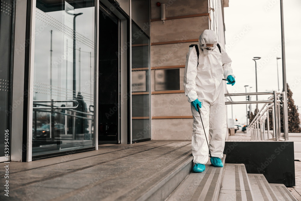 A male person in a white protective suit and mask disinfects the front door and stairs to the office building with chemicals. COVID-19 prevention sanitizing, corona, pandemic