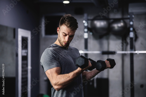 The smoldering look of an attractive and muscular man. A man in sportswear pumps his arm muscles and lifts dumbbells in a modern gym. Close up shot of a male person © dusanpetkovic1