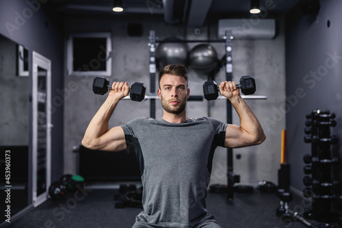 Muscular guy lifting dumbbell while sitting on a bench at the gym. A young athlete using dumbbells during a workout. Strong man under physical exertion pumping up bicep muscle with weight © dusanpetkovic1