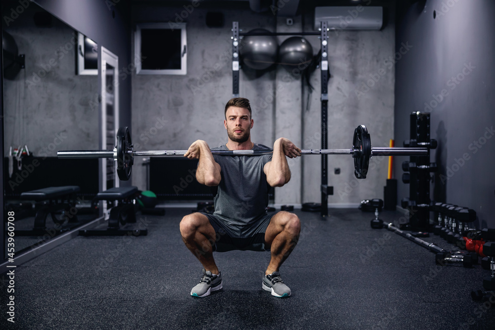Sports routine and active lifestyle. Front view of a young man in gray sportswear squatting with barbell in an indoor gym with equipment. Fitness body fell good and sexy, an attractive sports guy Sports routine and active lifestyle. Front view of a young man in gray sportswear squatting with barbell in an indoor gym with equipment. Fitness body fell good and sexy, an attractive sports guy