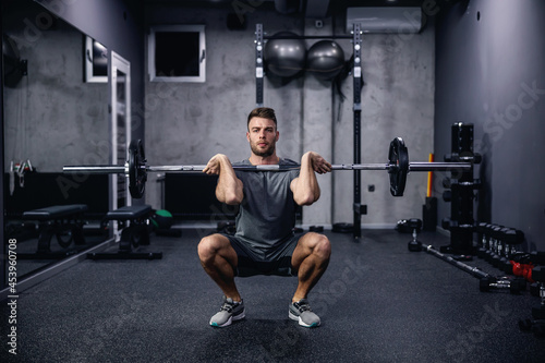 Sports routine and active lifestyle. Front view of a young man in gray sportswear squatting with barbell in an indoor gym with equipment. Fitness body fell good and sexy, an attractive sports guy