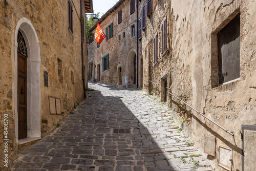 Fototapeta Naklejka Na Ścianę i Meble -  A narrow medieval stone paved street with a red waving flag in the tuscanian town of Montalcino