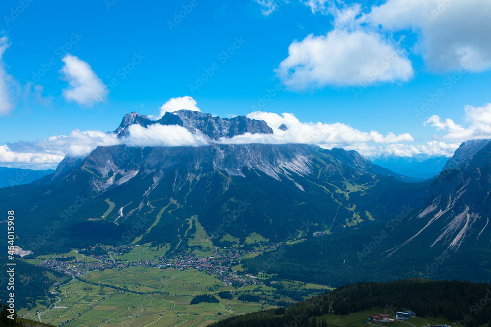Fototapeta premium Tyrolean Zugspitze Arena. Panoramic aerial view at the Zugspitze mountain with a big cumulus cloud. The highest mountain in Germany. Seen from Lermoos village. Tourism and vacations concept.