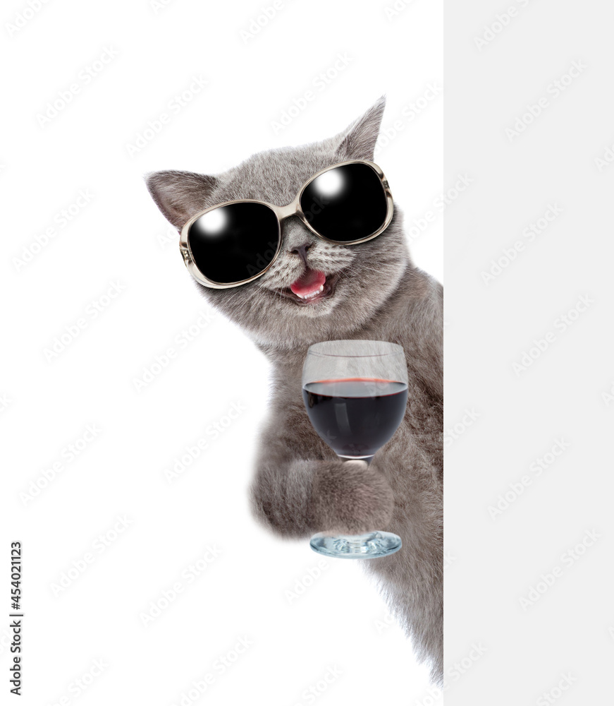 Happy cat wearing sunglasses holds glass of red wine and look from behind empty board. isolated on white background