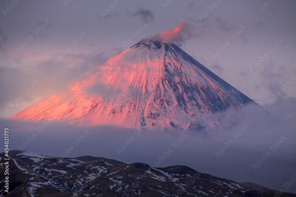 The cone of a high volcano covered with snow, colored by the red setting sun. Kamchatka volcano Klyuchevskoy