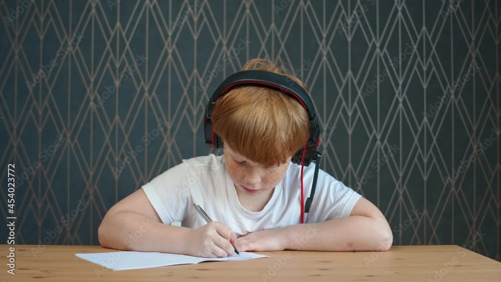 Homeschooling online by videocall, red-haired teen boy sitting at workplace in living room and uses a webcam to study in self-isolation at home, the boy writes in a notebook. Homeschooling online by videocall, red-haired teen boy sitting at workplace in living room and uses a webcam to study in self-isolation at home, the boy writes in a notebook.