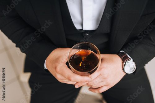 A man, a businessman in a black suit sits on a chair and holds a glass of alcohol, whiskey, brandy in his hands. Business photography.