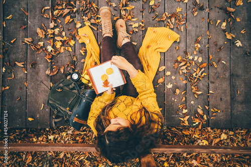 Woman with yellow scarf puts leaves between paper notebook pages sitting on wooden bridge in autumn forest close upper view. photo