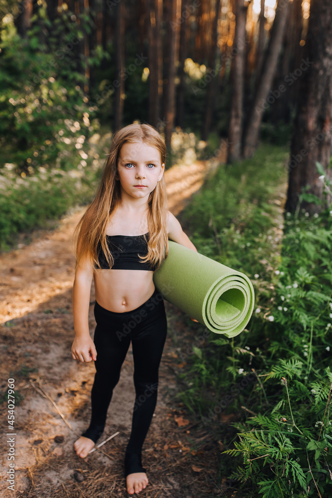 A beautiful preschool girl, a child athlete walks with a green rug in her hands for gymnastics and yoga in the forest at sunset. Sports photography. A beautiful preschool girl, a child athlete walks with a green rug in her hands for gymnastics and yoga in the forest at sunset. Sports photography.