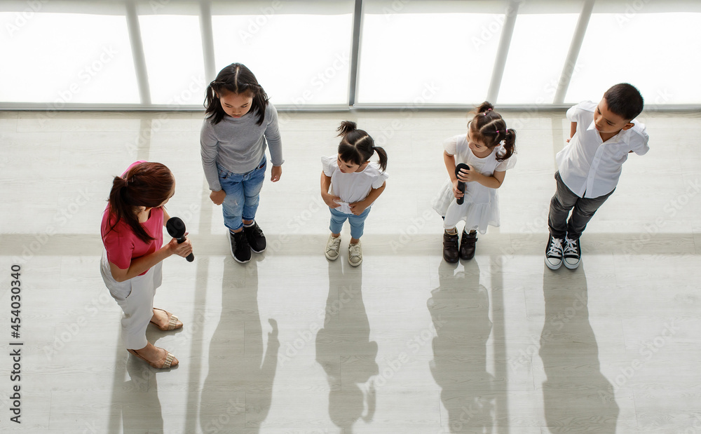 Top view shot of Asian female acting singing teacher hold microphone in hands teaching group of little young kindergarten boy and girls student learning to sing a song in music classroom studio
