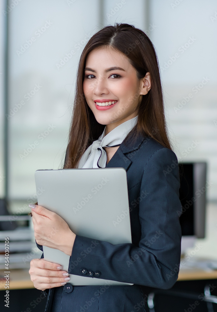 Portrait of young attractive Asian female office worker in formal business suits smiling at camera in office with blurry office as background Portrait of young attractive Asian female office worker in formal business suits smiling at camera in office with blurry office as background