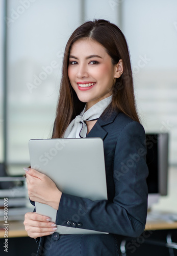 Portrait of young attractive Asian female office worker in formal business suits smiling at camera in office with blurry office as background