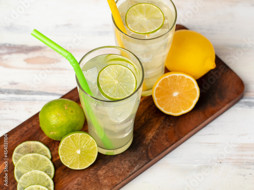 Refreshing glasses of cold fruit juice, ice lime and lemon juice, decorating with fresh lime and lemon slice on wooden chopping board on white table. Yellow and green straw are next to it