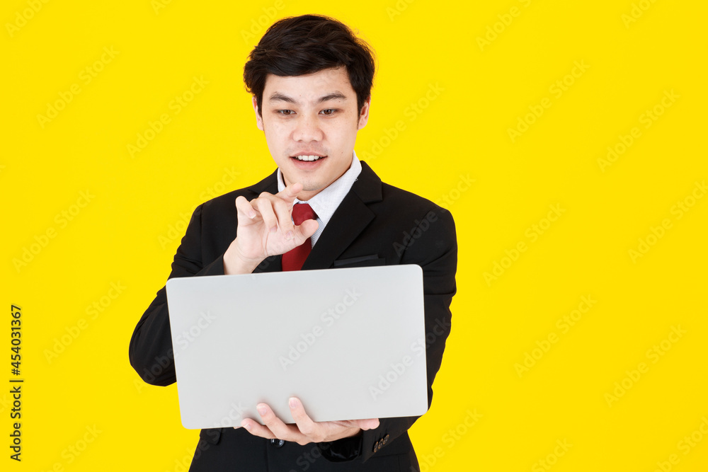 Horizontal portrait shot of young adult Asian man in a black long sleeve suit holding a closed laptop while trying to touch something in empty space in air isolated with yellow background in studio Horizontal portrait shot of young adult Asian man in a black long sleeve suit holding a closed laptop while trying to touch something in empty space in air isolated with yellow background in studio