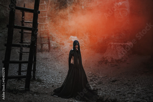 Young woman in witch stands in black dress in dark and foggy forest.