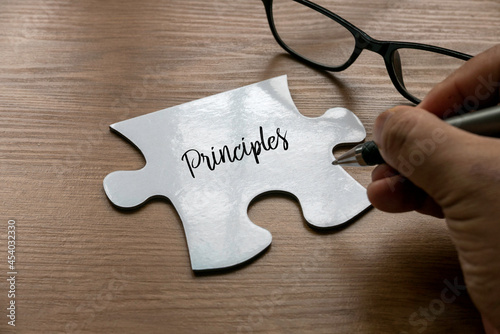 Selective focus of a hand writing word ' Principles ' on a a piece of white jigsaw puzzle with a glasses on wooden background. photo