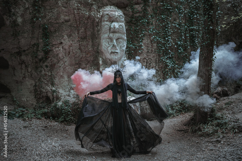 Young woman in witch stands in black dress and crown on her head in dark and foggy forest.