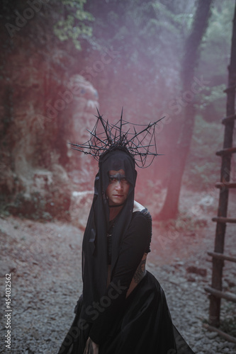Young woman in witch stands in black dress and crown on her head in dark and foggy forest.