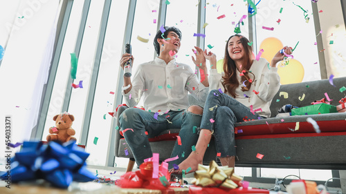 Lovely young Asian couples enjoy celebrating new year festival by playing colorful paper flares in living room decorated with gift box  fancy balloon for funny party on beautiful day in winter