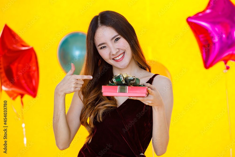 Asian pretty girl in sexy dress standing smiling holding and pointing green ribbon present gift red box in front colorful star helium air balloons on yellow background in surprise birthday party Asian pretty girl in sexy dress standing smiling holding and pointing green ribbon present gift red box in front colorful star helium air balloons on yellow background in surprise birthday party