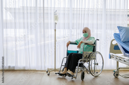 Portrait of ill Muslim wearing patient gown and face mask sitting on a wheelchair with saline fluid bag attach in hospital. Covid 19 pandemic concept