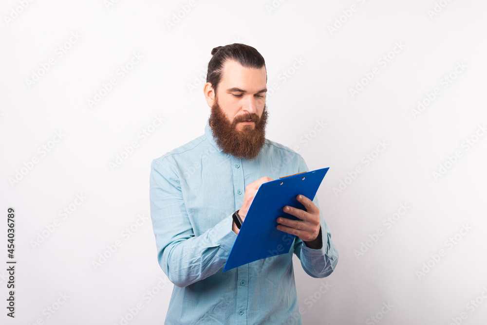 Serious bearded man is writing something on a paper holder board while standing over white background. Serious bearded man is writing something on a paper holder board while standing over white background.