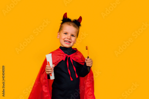 One little toddler boy in a carnival costume with toothpaste and an orange brush for Halloween is isolated on a yellow background. Medicine, dental hygiene, holidays concept.