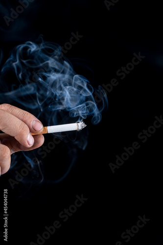 Black background. the hand of a man with roughened skin holds a cigarette. blue smoke is coming from it. close-up.