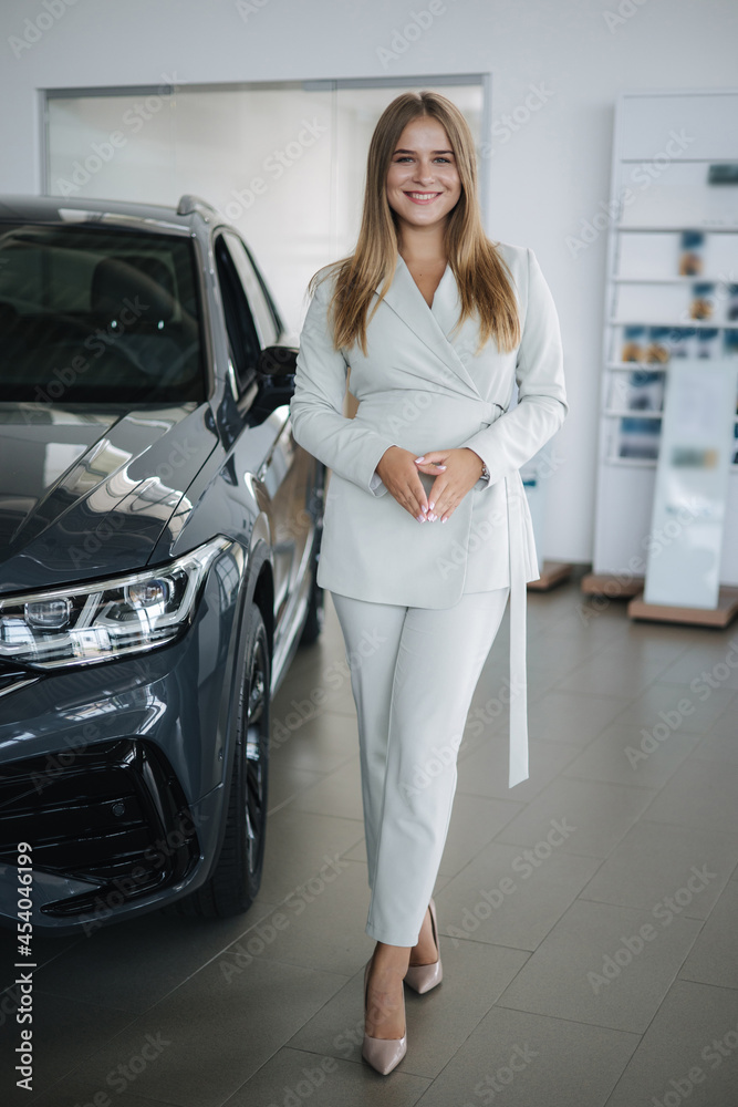 Portrait of gorgeous woman in showroom. Beautiful salesperson standing by car. Blond hair female after buying car