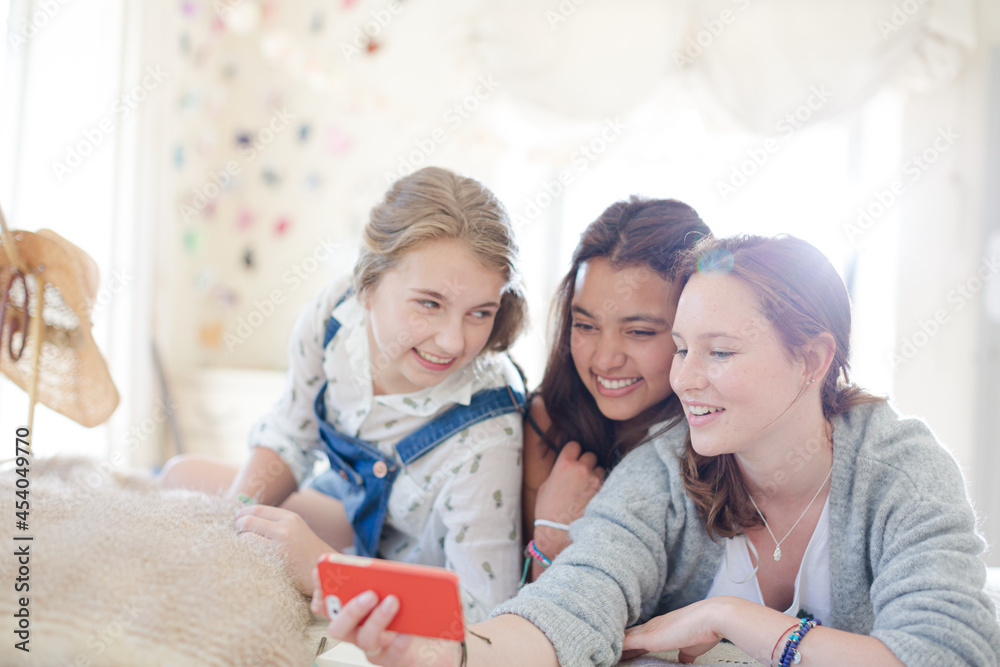 Three teenage girls using smart phone together while lying on bed in bedroom