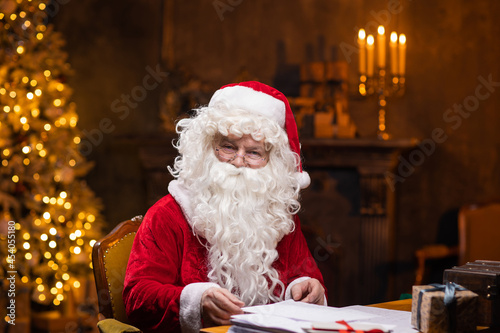 Workplace of Santa Claus. Cheerful Santa is working while sitting at the table. Fireplace and Christmas Tree in the background. Christmas concept.
