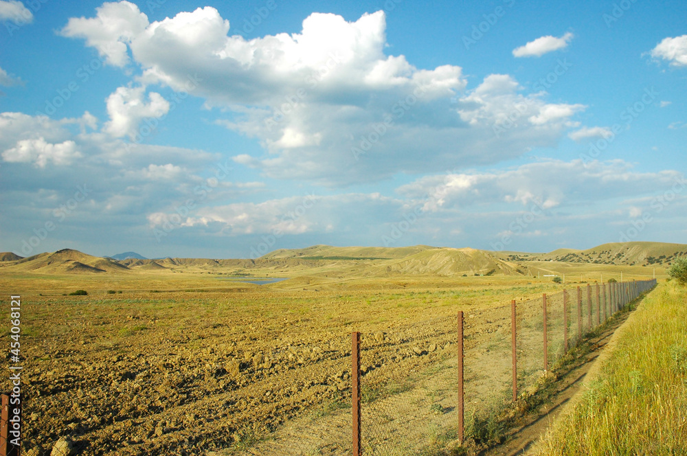 A field fenced with a chain-link fence. Sandy mountains and a lake on the horizon. The road to Cape Meganom. Crimea.