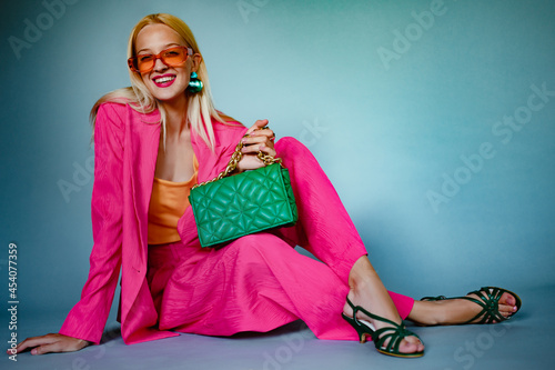 Happy smiling fashionable woman wearing trendy pink fuchsia color suit, orange sunglasses, strappy sandals, with green quilted casette leather bag. Model posing on blue background. Copy, empty space 