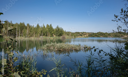 Fototapeta Naklejka Na Ścianę i Meble -  A lake originated from an abandoned and flooded quarry overgrown with trees and lush grass. Reed islands are scattered throughout the water. Summer in the daytime.