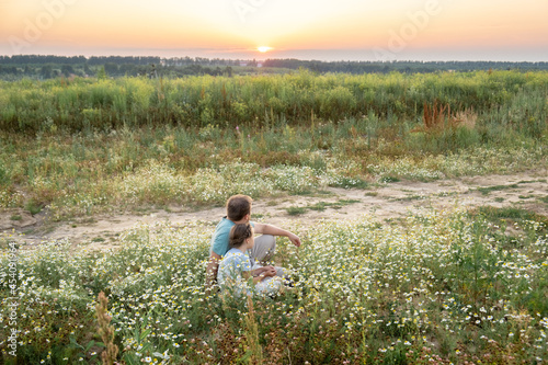 a father and a little daughter sit side by side on the grass in a chamomile field on a sunny summer day and look into the distance at the sunset. General plan