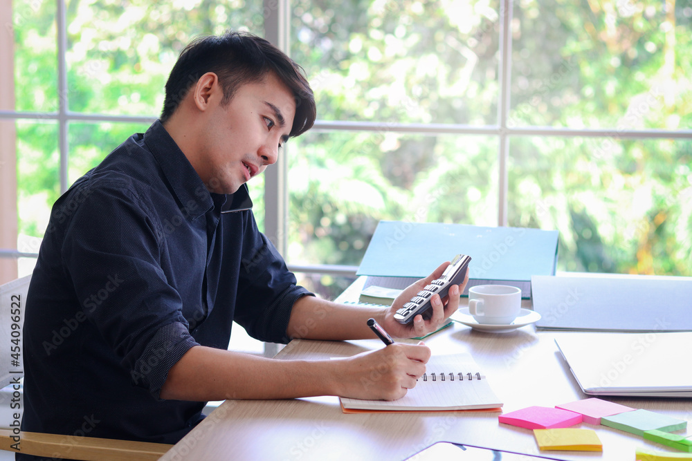 Busy businessman in black shirt working on office desk, taking notes while talking with customer on mobile phone, and the other hand holding a calculator, writing information data on paper. Busy businessman in black shirt working on office desk, taking notes while talking with customer on mobile phone, and the other hand holding a calculator, writing information data on paper.