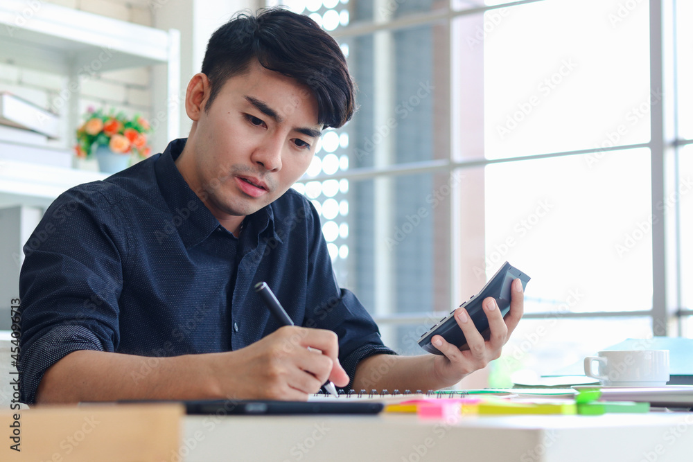 Asian young handsome businessman in black shirt working on office desk, taking notes while the holding a calculator on other hand, writing financial information data on paper for analysis. Asian young handsome businessman in black shirt working on office desk, taking notes while the holding a calculator on other hand, writing financial information data on paper for analysis.